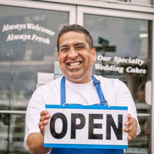 Latino man standing in front of his store, smiling holding an "Open" sign